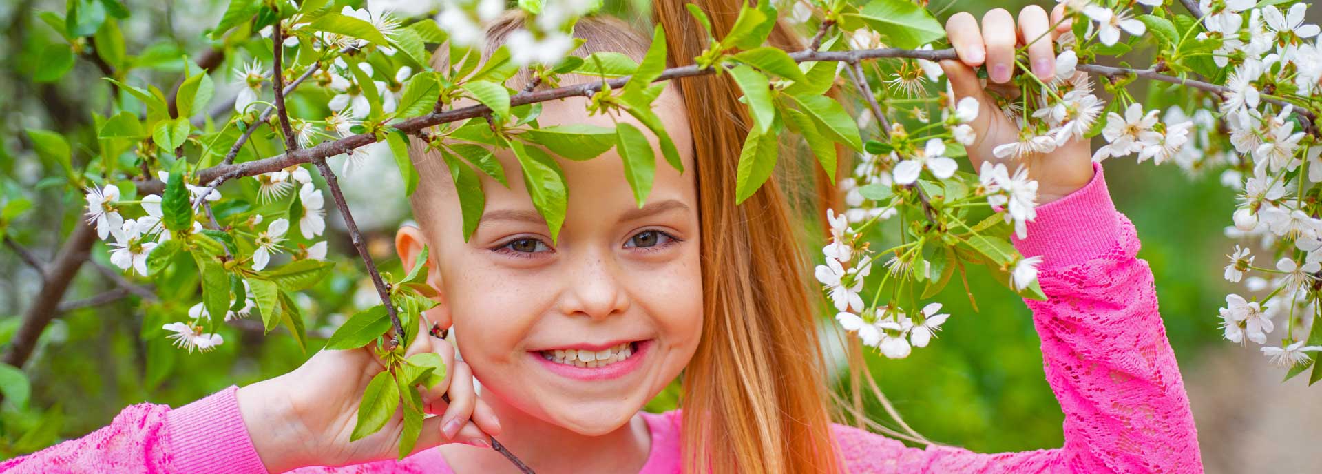 Close up of young girl smiling at camera standing among flowering tree branches.