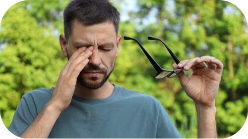 Man in forested area removing glasses and rubbing his right eye.